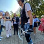 Słoneczny dzień, niebieskie niebo, deptak w parku. Na deptaku stoją w rozsypce ludzie ubrani w letnie ubrania. Na pierwszym planie młody mężczyzna ubrany w symulator starości przemieszcza się przy wsparciu balkonika. Nastrój zdjęcia pogodny.