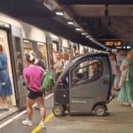 Te sam peron metra. Widok wzdłuż pociągu. Do wagonów wsiadają różni ludzie. wśród nich widoczny szary skuter.