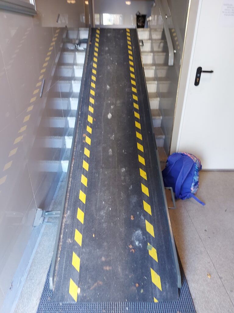 Ta sama klatka schodowa. Pochylnia jest tym razem rozłożona, Jest to gruba, czarna blacha, z odbojnikami wzdłuż całej długości po obu stronach. Pochylnia jest koloru czarnego, wzdłuż jej biegu na nawierzchni zaznaczono przerywaną, żółtą linią dwie proste. Całość jest bardzo stroma i zajmuje cała szerokość użytkową schodów. Przed schodami, koło pochylni stoi niebieski plecaczek.