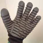 Na jasny tle dłoń ubrana w rękawice. Widzimy wewnętrzną stronę dłoni – tu rękawica jest gumowa z licznymi dodatkowymi utwardzeniami. Na nadgarstku osoby pozującej widać zielony pasek od zegarka i 2 bransoletki z kolorowych, okrągłych, szklanych koralików.