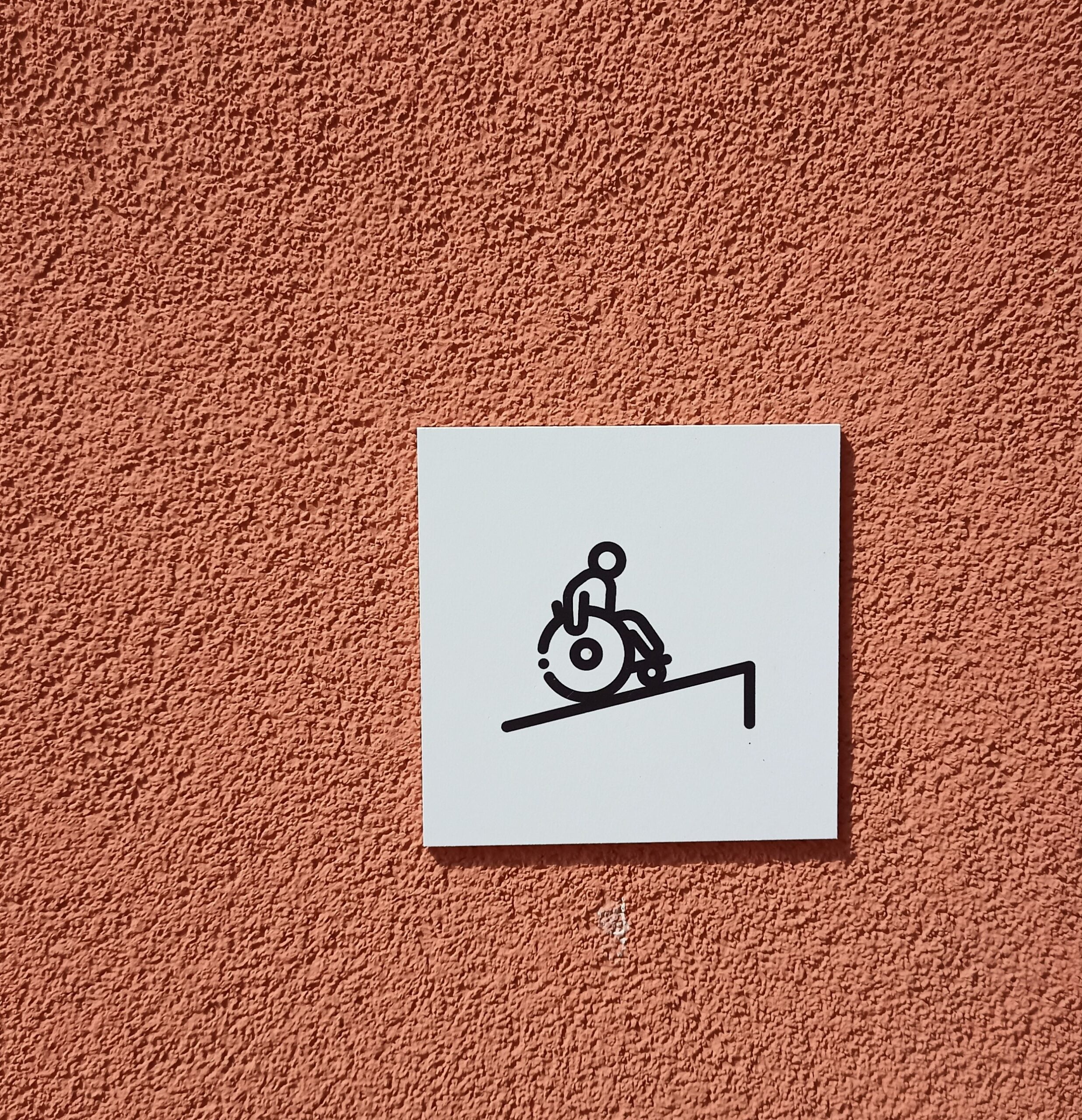 Na ceglastej w kolorze ścianie jasnoszara, kwadratowa tabliczka z czarnym piktogramem osoby na wózku podjeżdżającej na pochylnię.