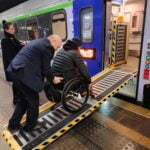 Peron na dworcu kolejowym. Przy nim pociąg z otwartymi drzwiami, między wejściem do wagonu i peronem rozłożona rampa przenośna. Konduktor pomaga pokonać rampę mężczyźnie na wózku.