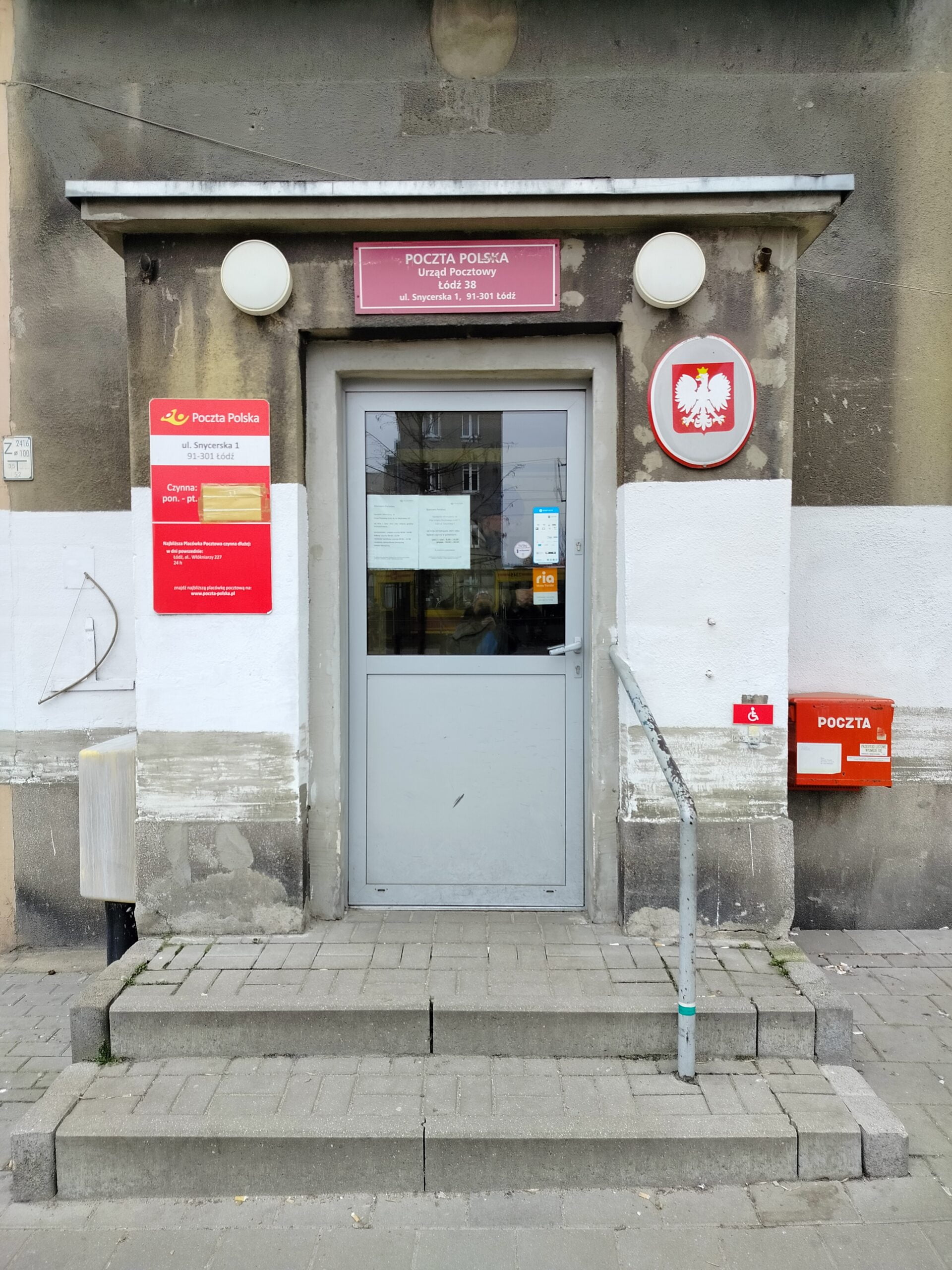 Widok z przodu na drzwi wejściowe do budynku. do drzwi prowadzą 2 stopnie z poręczą po prawej stronie. Nad prawą krawędzią schodów, za pochwytem widoczny biały piktogram osoby na wózku na czerwonym tle. Na drzwiami tablica informująca że jest to placówka poczty polskiej, po lewej stronie drzwi tablica z godzinami otwarcia, po prawej stronie drzwi godło polskie.
