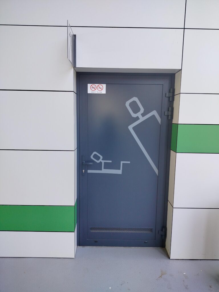 Widok frontalny na grafitowe drzwi. Na nich jasnoszare nieoczywiste rysunki. Jeden z nich to jakby kwadratowate dziecko na desce a drugi to człowiek robiący a kuku zza winkla.