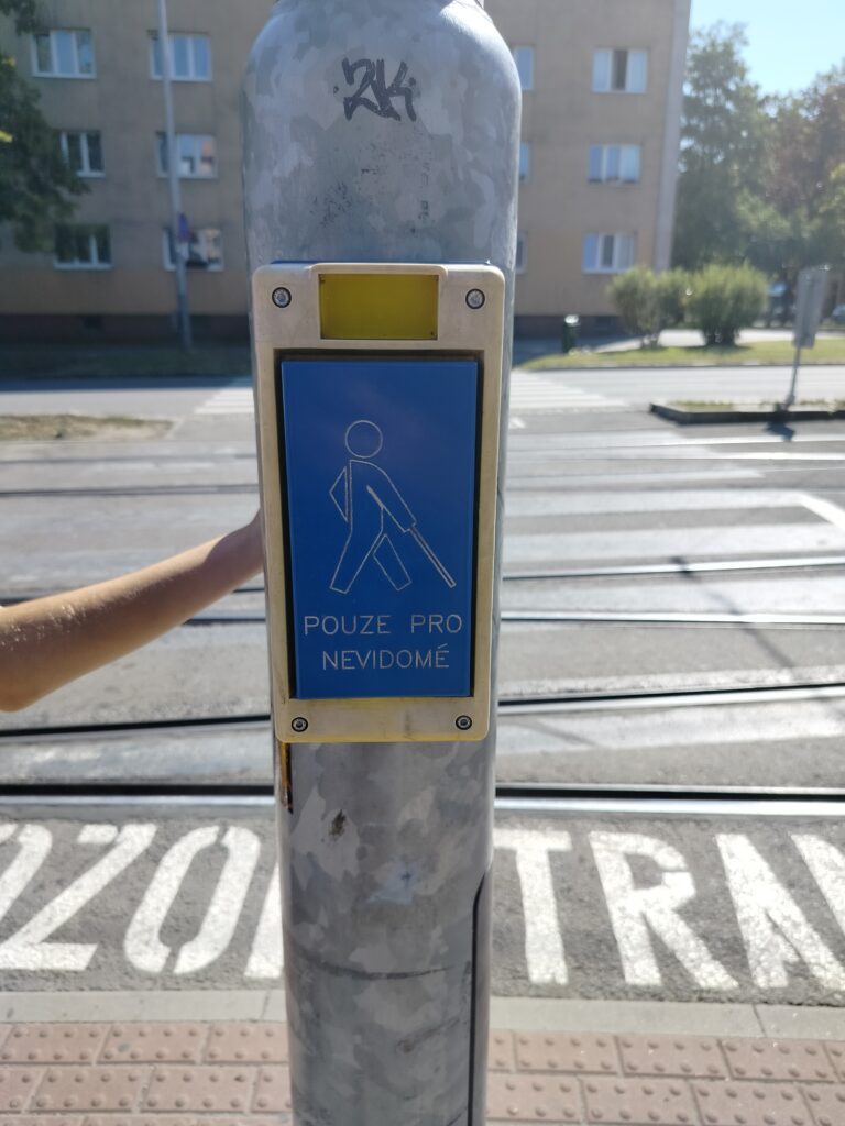 Widok na przejście przez jezdnię i tory tramwajowe. Na planie głównym słup z przyciskiem. W żółtej obudowie duży niebieski przycisk z piktogramem osoby z laską dla niewidomych oraz napis POUZE PRO NEVIDOME