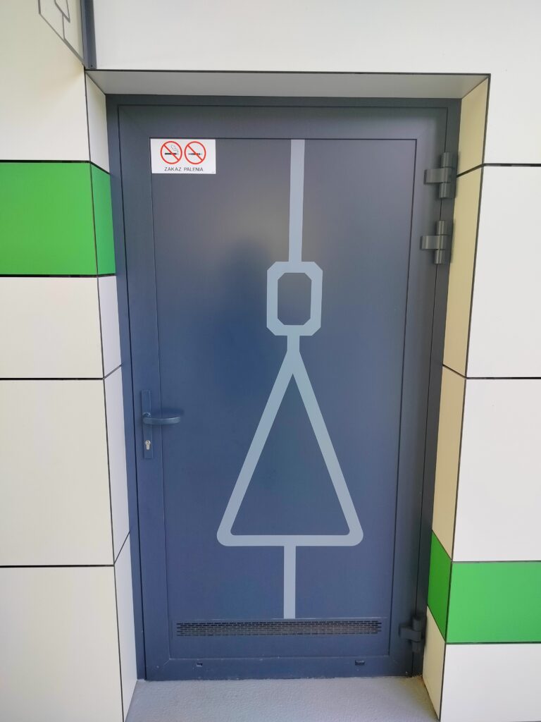 Widok od frontu na grafitowe drzwi, na nich jasnoszary piktogram nawleczonej na linię sylwetki ludzkiej - w miejscu głowy ośmiokąt foremny a w miejscu tułowia trójkąt równoramienny o podstawie równoległej do podłogi.