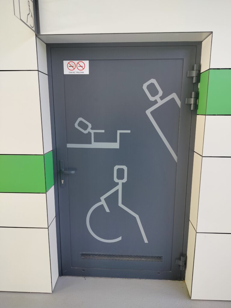 Widok od frontu na grafitowe drzwi, na nich 3 jasnoszare piktogramy. Pierwszy z nieznacznie udziwnionych piktogramem osoby na wózku-osoba ma ośmiokątną głowę a koło wózka jest niedomknięte. Drugi to kwardarowatych kształtów dziecko położone na poziomej desce, trzeci to osoba robiąca a kuku zza winkla.