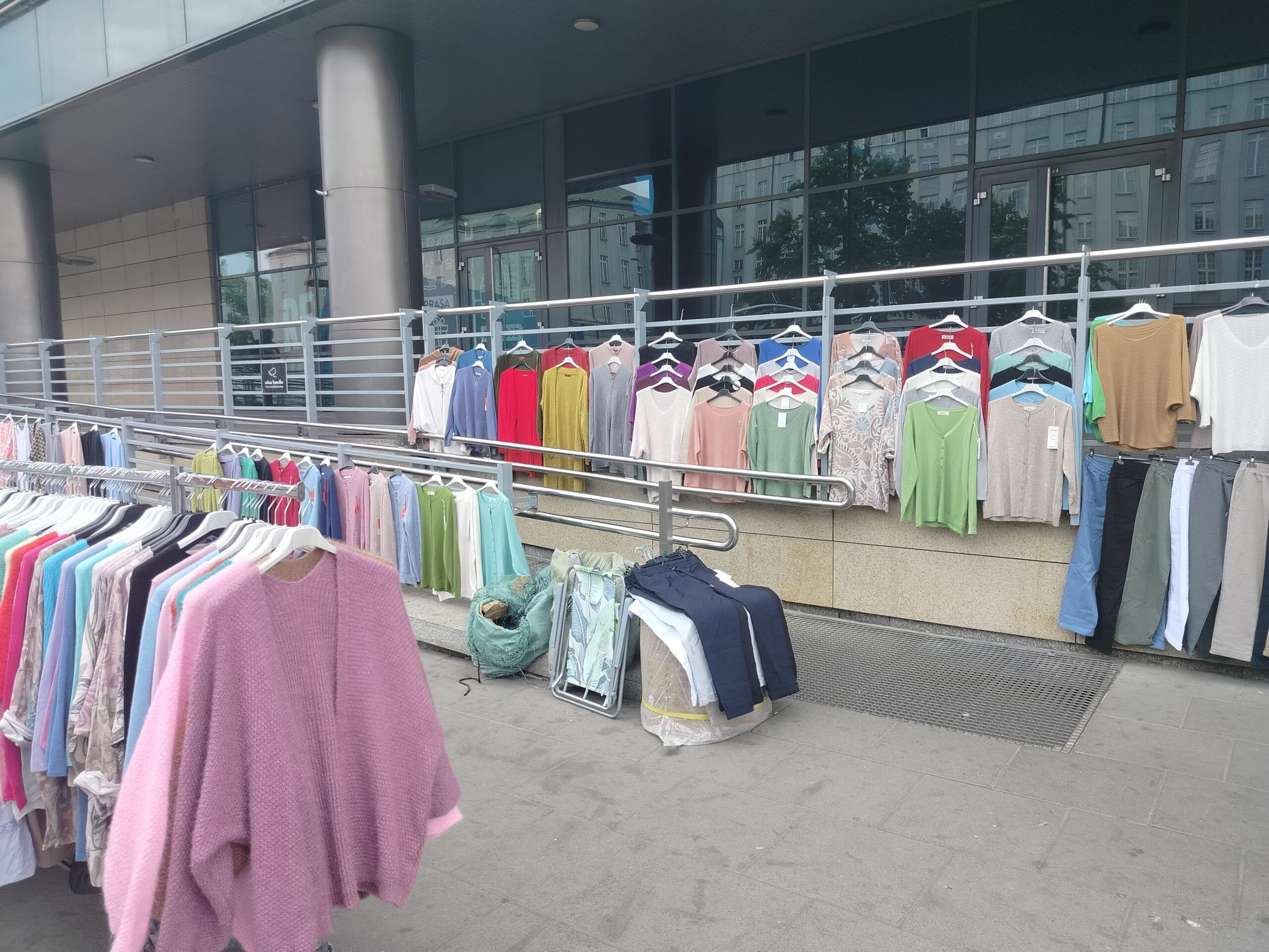 Widok na pochylnię z boku na jej pochwytach od strony chodnik wiszą kolorowe bluzki i spodnie na plastikowych wieszakach. Z przodu kadru wieszak sklepowy na wieszaki a na nich kolejne ubrania.